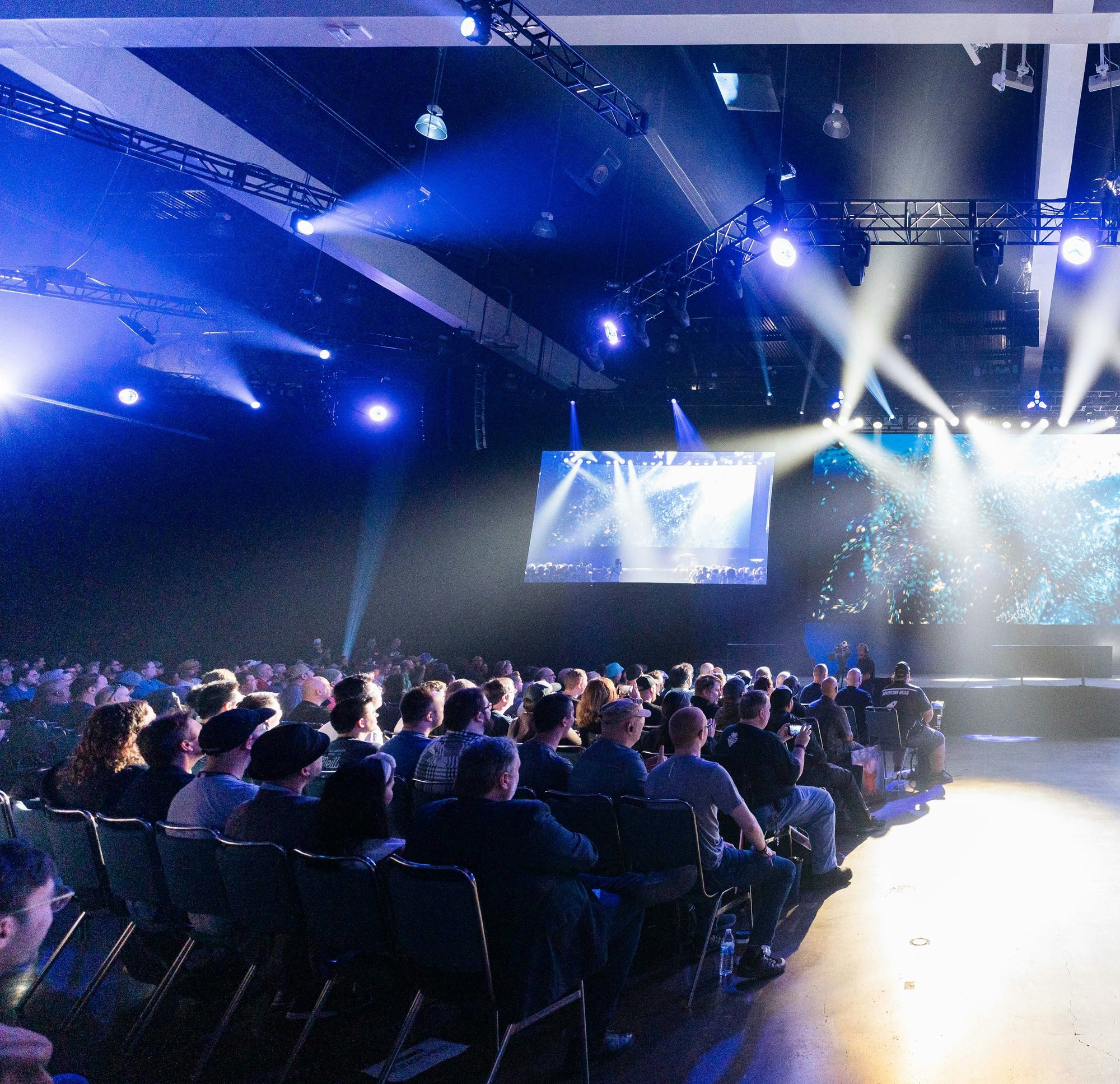 A summit hall under blue stage lighting with a seated audience facing the stage screens